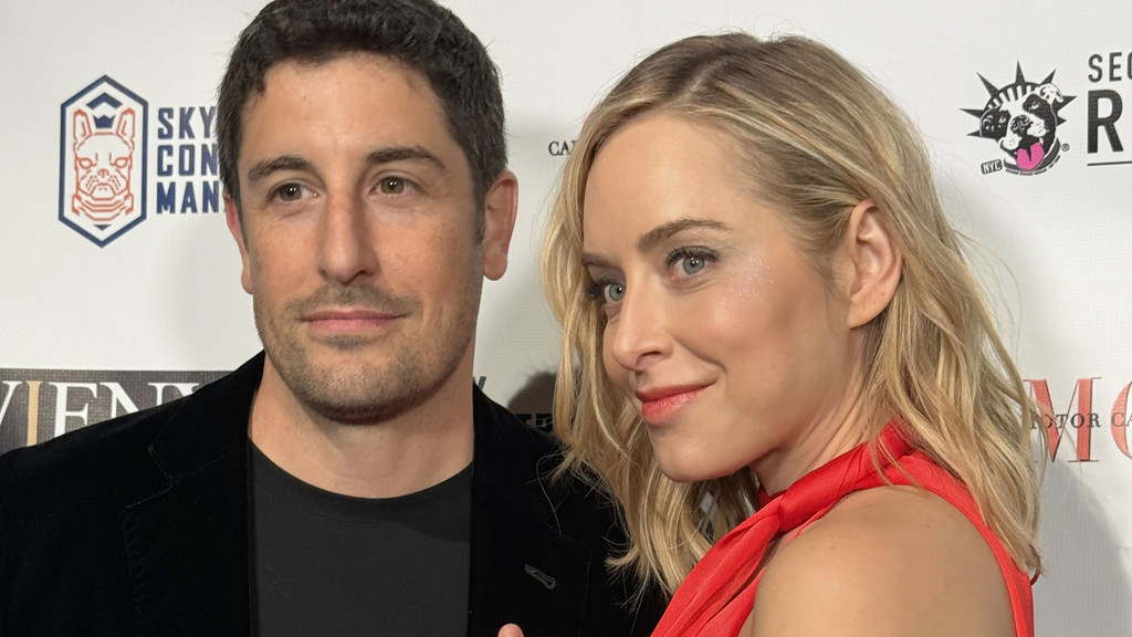 Jason Biggs, in a black jacket, and Jenny Mollen, in a red dress, pose smiling at an event against a white backdrop with various logos.