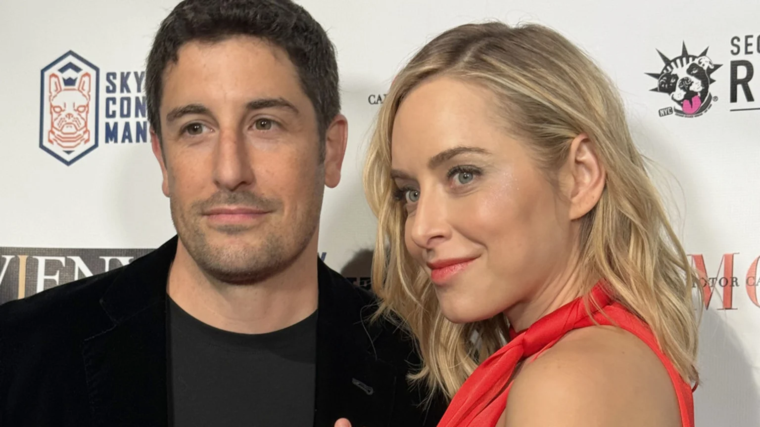 Jason Biggs, in a black jacket, and Jenny Mollen, in a red dress, pose smiling at an event against a white backdrop with various logos.
