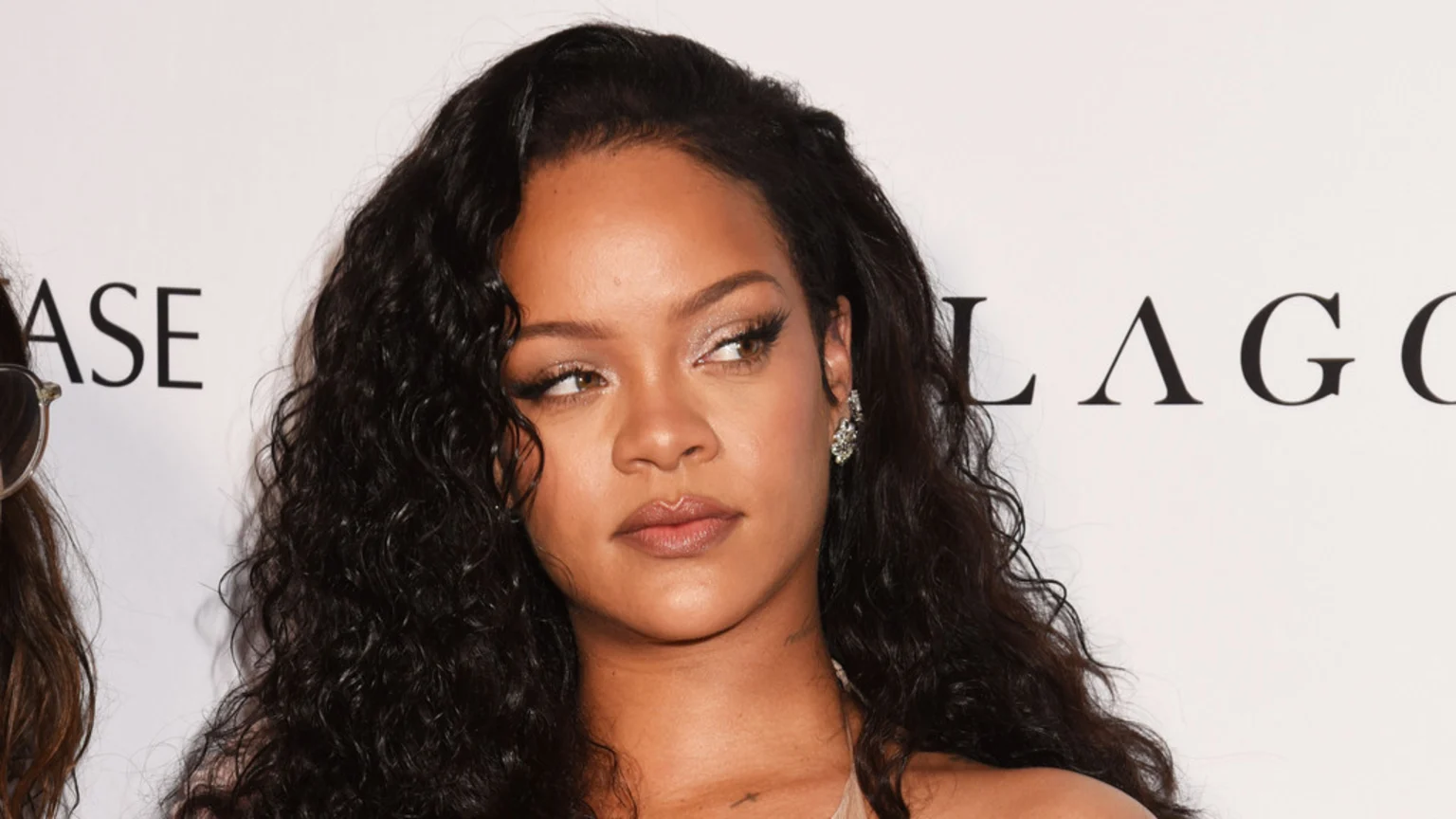 Close-up of Rihanna with long, dark curly hair, wearing an earring, looking to the right against a white background with black text.