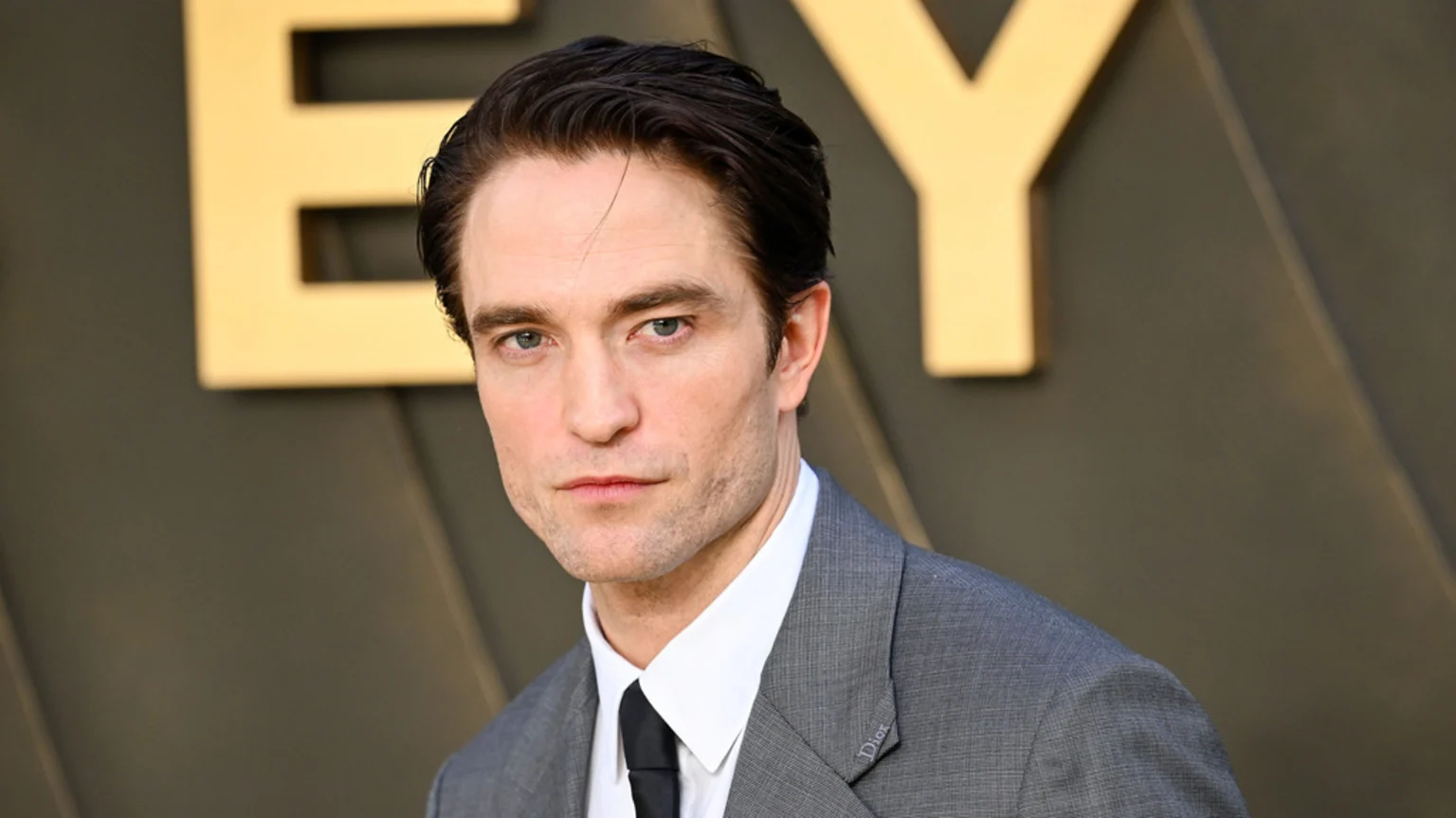 A close-up of actor Robert Pattinson, wearing a grey suit with a white shirt and black tie, looking directly at the camera with a serious expression.