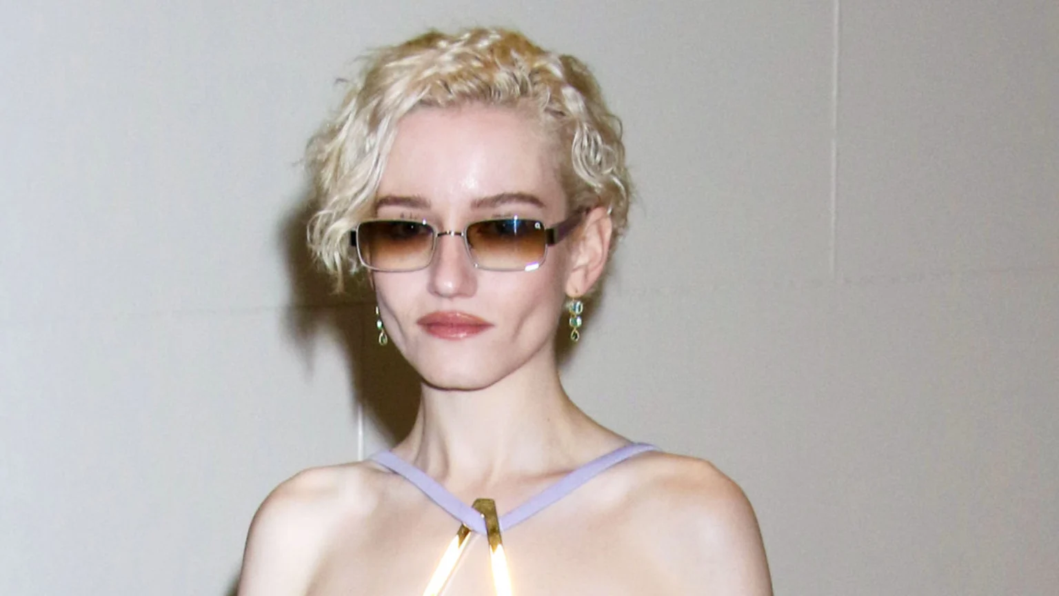 Julia Garner, with short curly blonde hair and dimples, wears rectangular gradient sunglasses, green beaded earrings, and a light purple top with a gold detail, looking towards the camera.