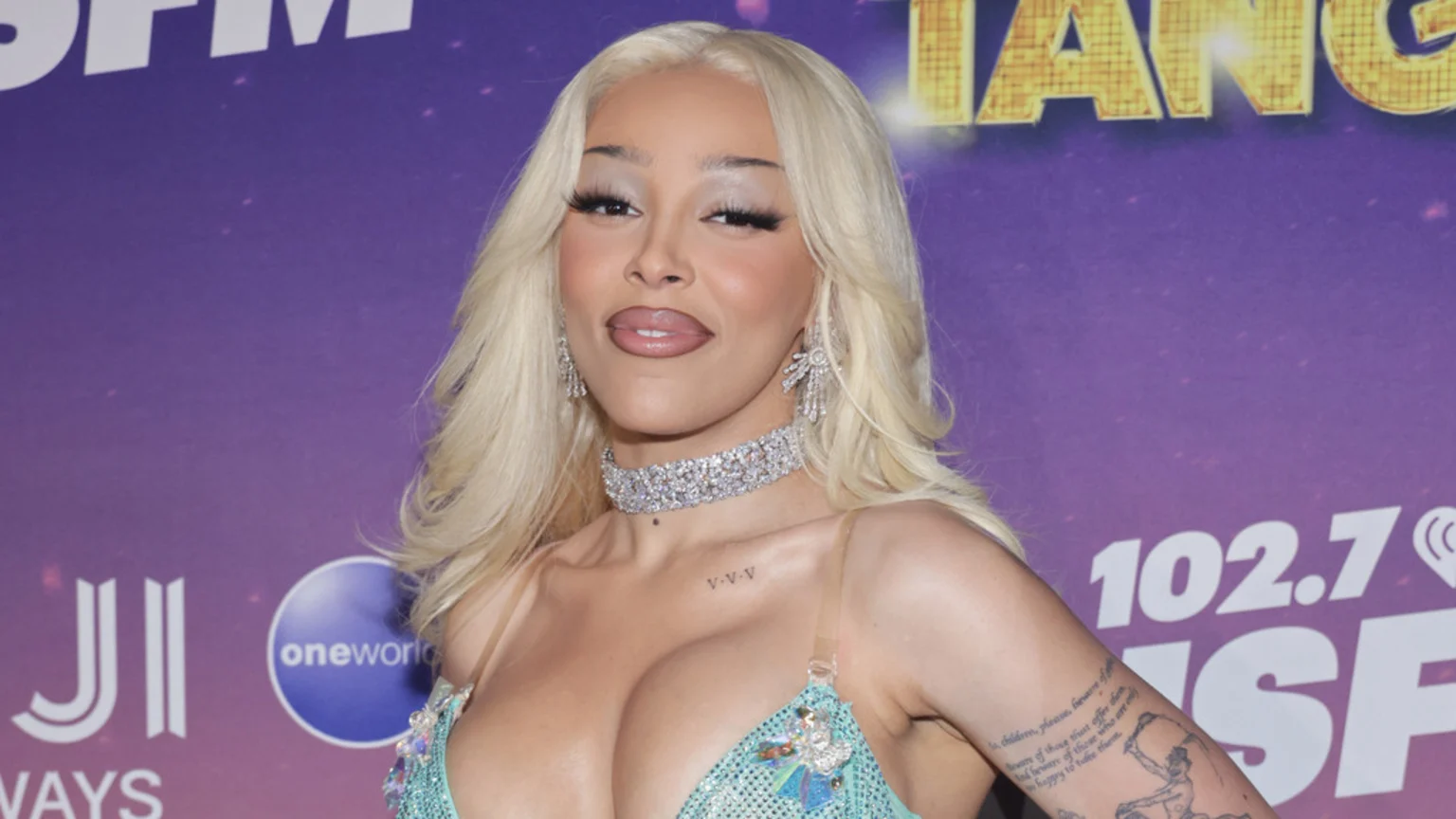 Doja Cat, with long blonde hair and dramatic makeup, wears a sparkling diamond choker, matching earrings, and a teal sequined top while posing in front of a purple background with various logos.