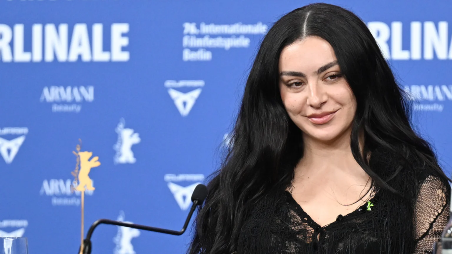 A woman with long dark hair and a faint smile wears a black top, sitting at a table with a microphone and a golden bear figure, against a blue Berlinale logo background.