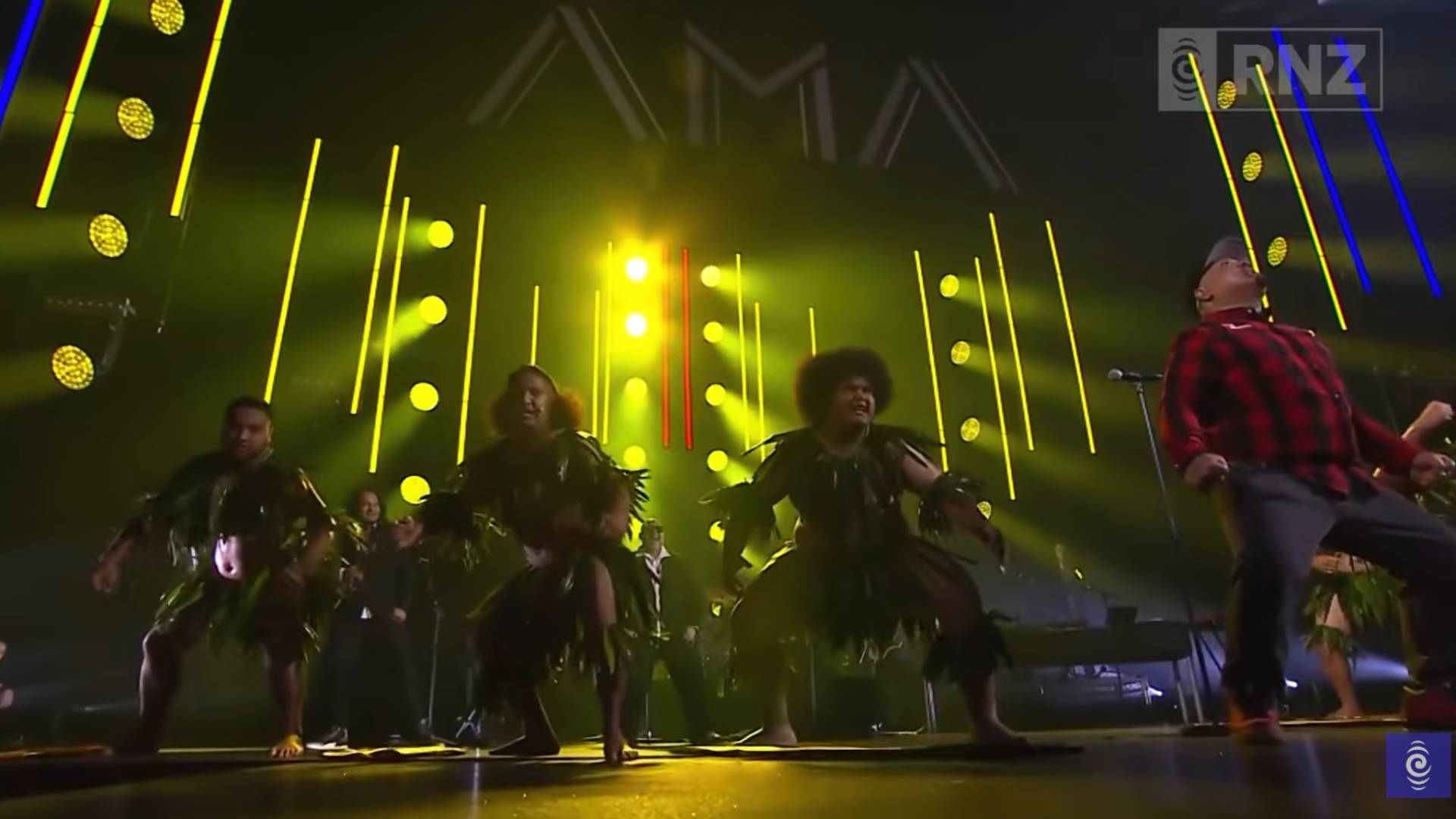 Two dancers in leafy traditional costumes perform on a brightly lit stage with yellow lights, with a man in a red plaid shirt performing on the right.