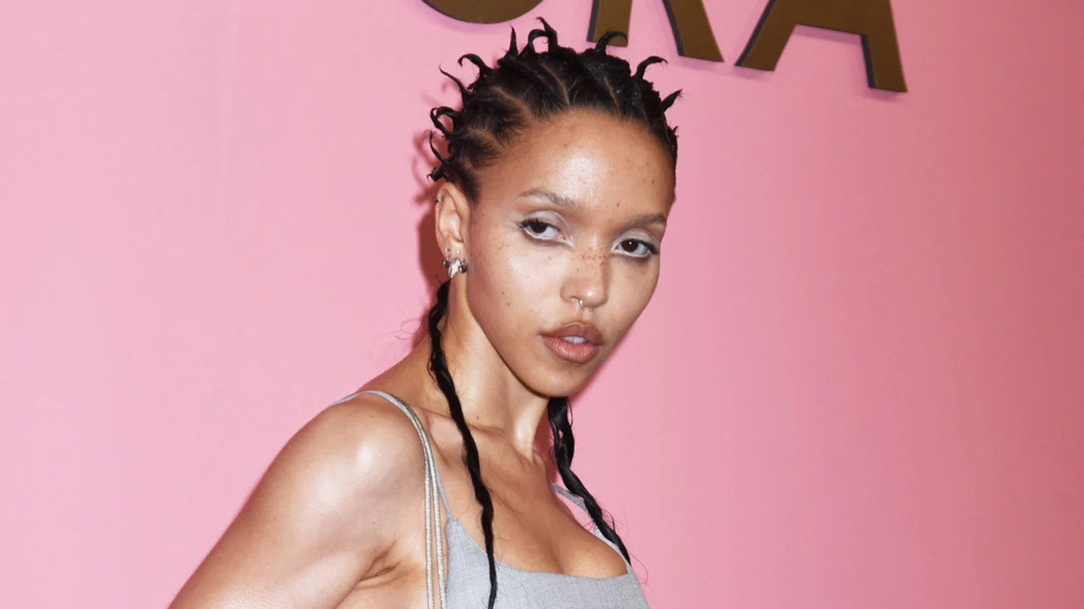 A woman with braided hair featuring upward-sticking twists and long hanging braids, white eyeliner, facial and ear piercings, wearing a light grey top, poses against a pink background with a dark letter.