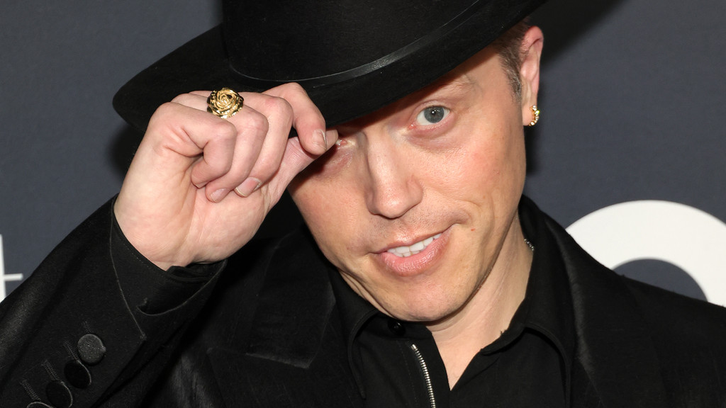A man in a black hat and jacket with a gold rose ring and earring looks at the camera while tipping his hat.