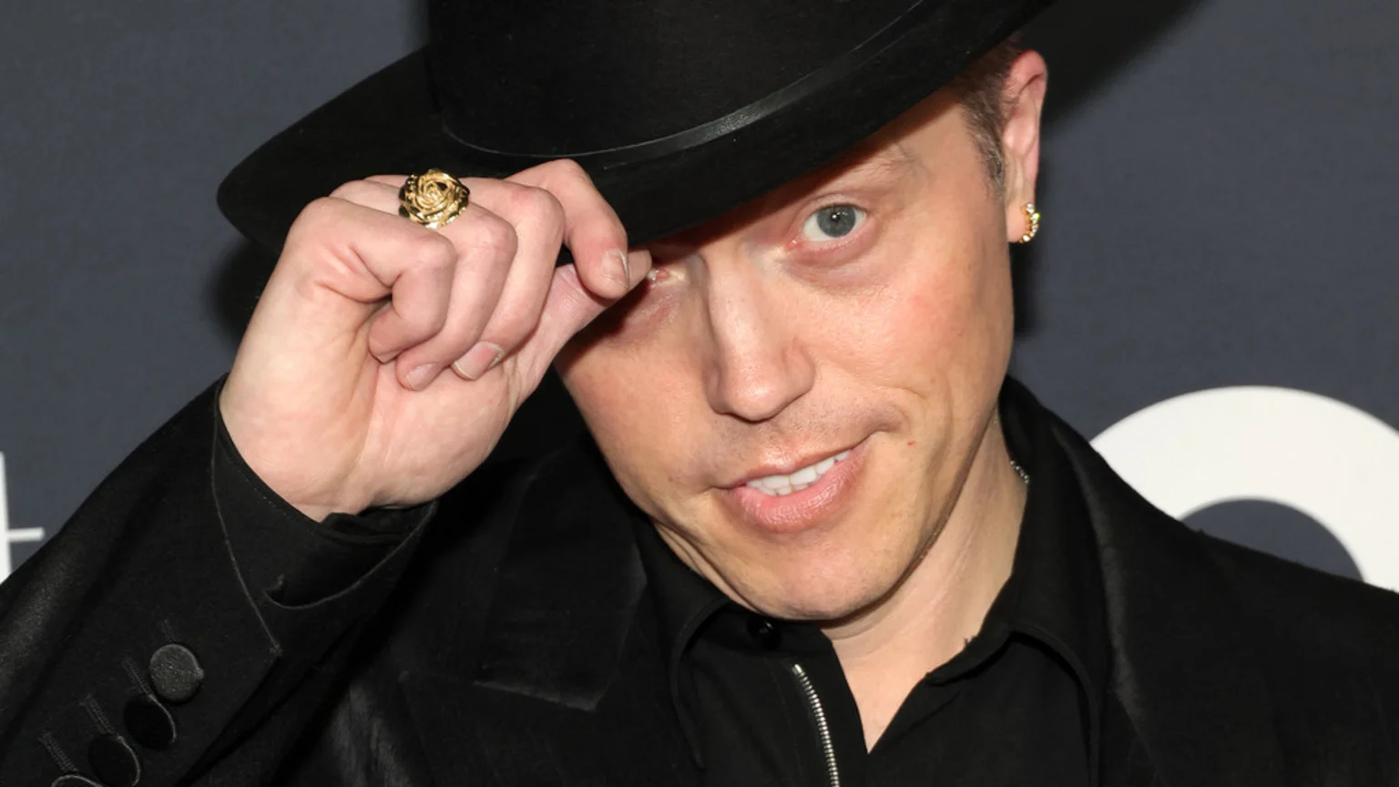 A man in a black hat and jacket with a gold rose ring and earring looks at the camera while tipping his hat.