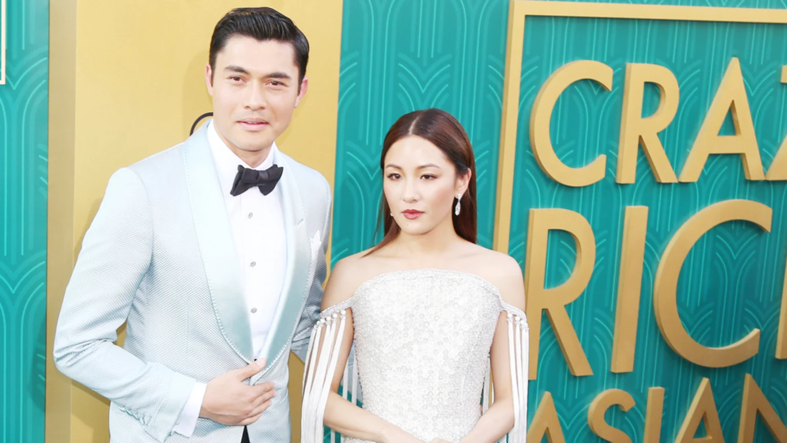 Henry Golding in a light blue tuxedo and a woman in a white beaded dress stand in front of a teal and gold "Crazy Rich Asians" backdrop.