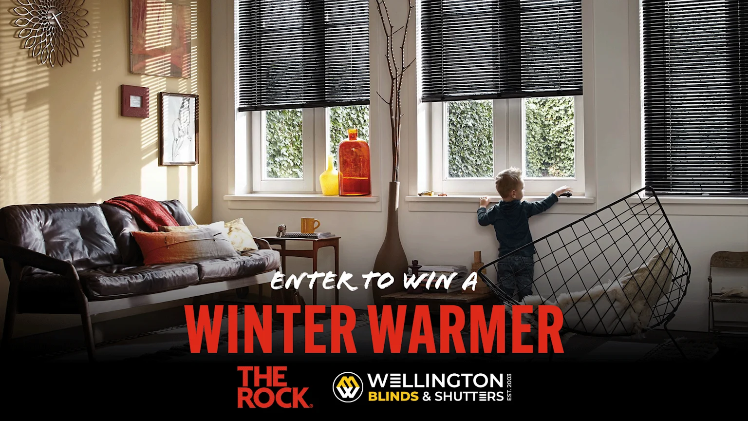 An advertisement showing a living room with black window blinds, a dark leather couch, a child looking out a window, and text 'ENTER TO WIN A WINTER WARMER' with The Rock and Wellington Blinds & Shutters logos.