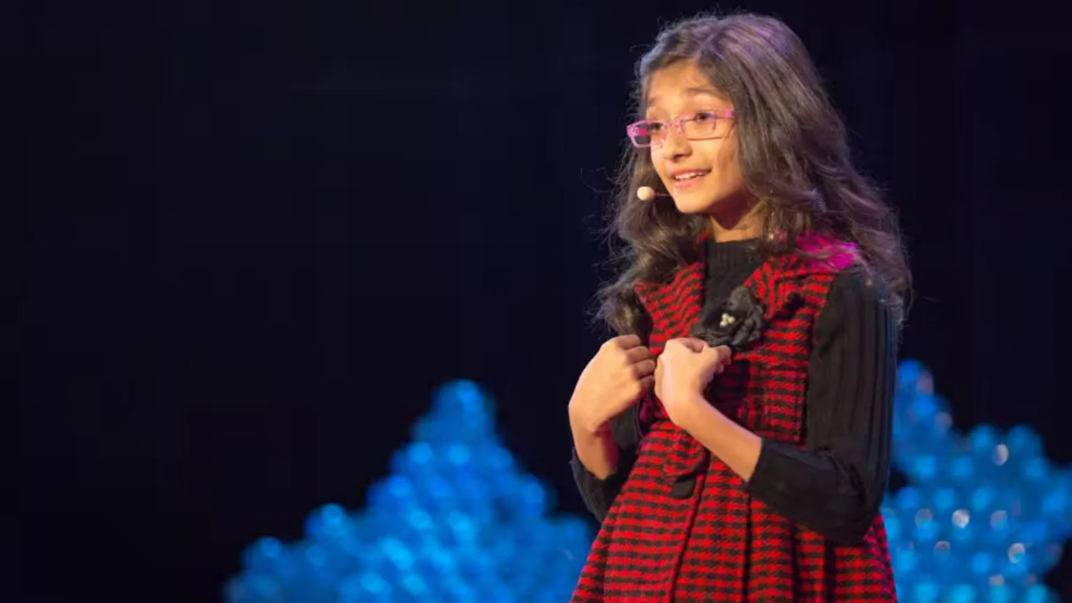 A young girl with long dark hair, pink glasses, and a red and black plaid outfit smiles on a stage, her hands clasped, with blurred blue lights in the background.