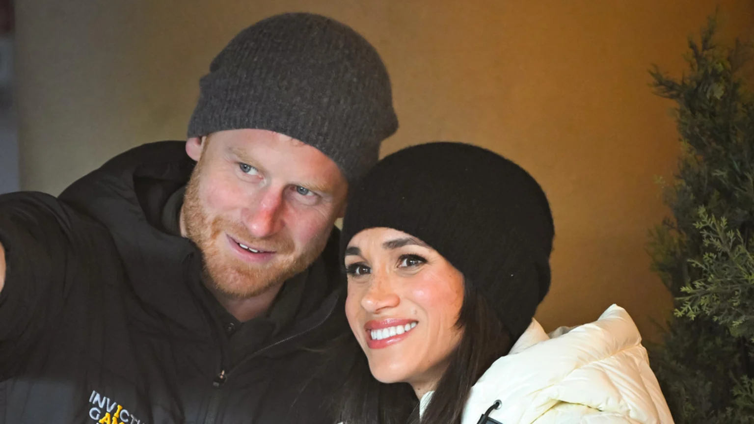 Prince Harry, in a grey beanie and black jacket, and Meghan Markle, in a black beanie and white puffer jacket, smile while taking a selfie together.