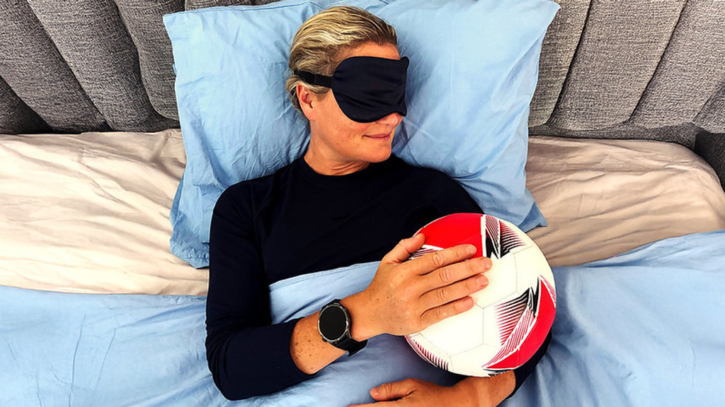 A person wearing a sleep mask lies in bed under blue covers, cuddling a red and white soccer ball.