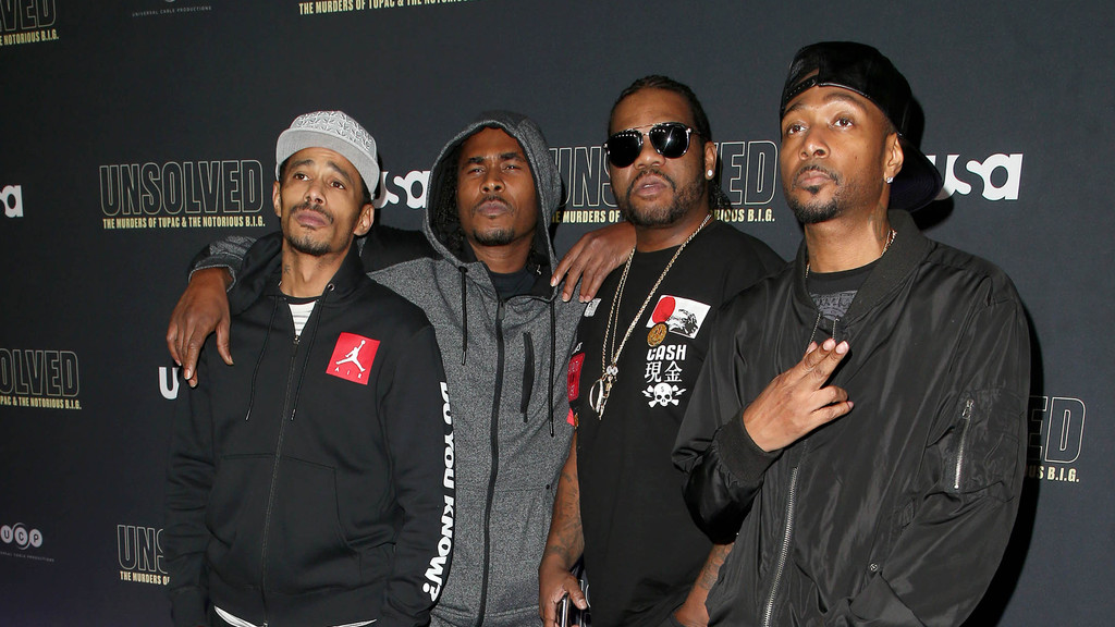 Four men stand together, one in a grey cap, one in a grey hoodie, one in sunglasses and a black tee, and one in a black bomber jacket, against a dark "UNSOLVED" backdrop.