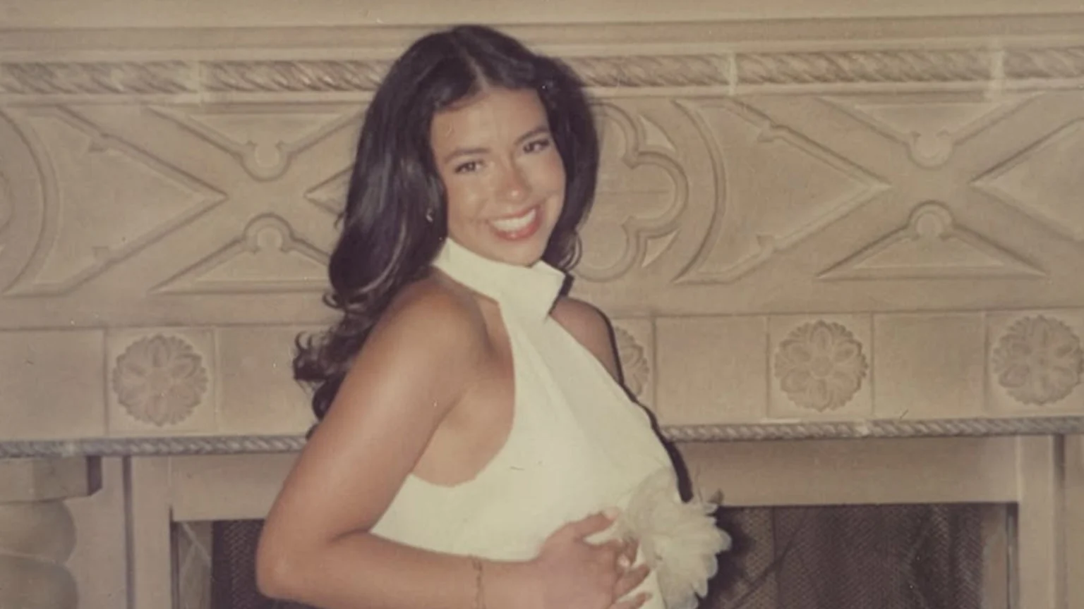 A smiling woman with long dark hair wears a light-colored halter top and holds her stomach, standing in front of a carved stone fireplace.