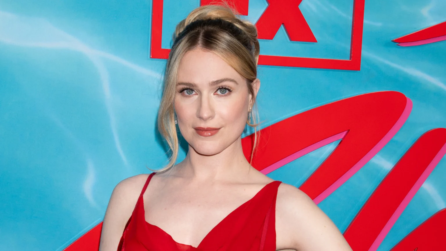 Evan Rachel Wood, a blonde woman in a red dress with her hair pulled up, smiles slightly against a light blue background featuring red letters.