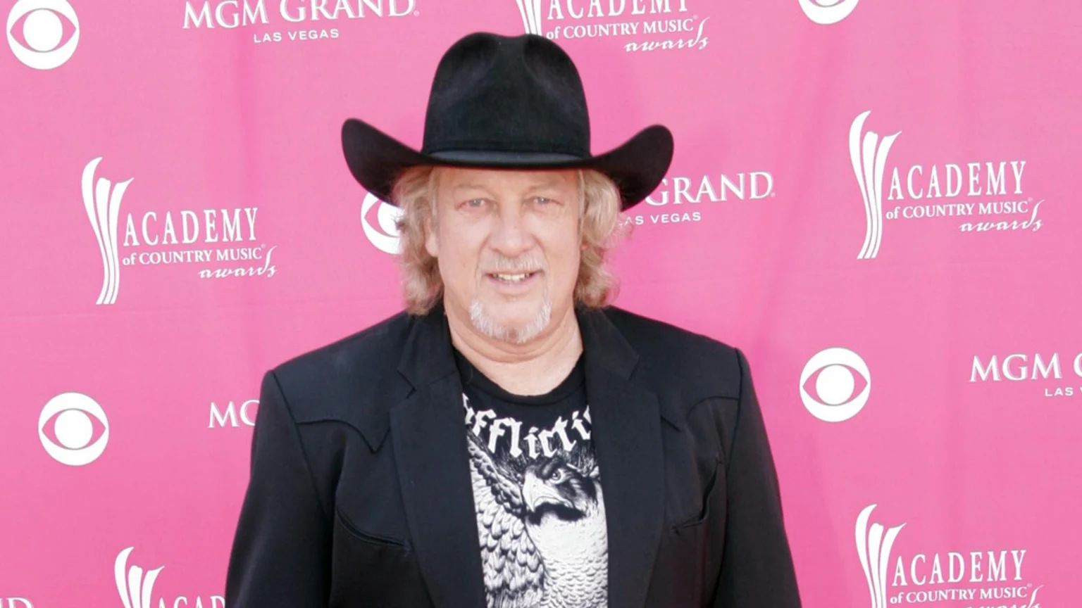 Country singer John Anderson, wearing a black cowboy hat, blazer, and graphic tee, smiles in front of a pink Academy of Country Music Awards backdrop.