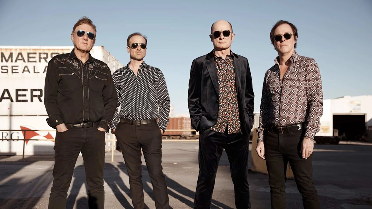 Four men wearing sunglasses and patterned shirts stand outdoors in front of white shipping containers and industrial buildings.