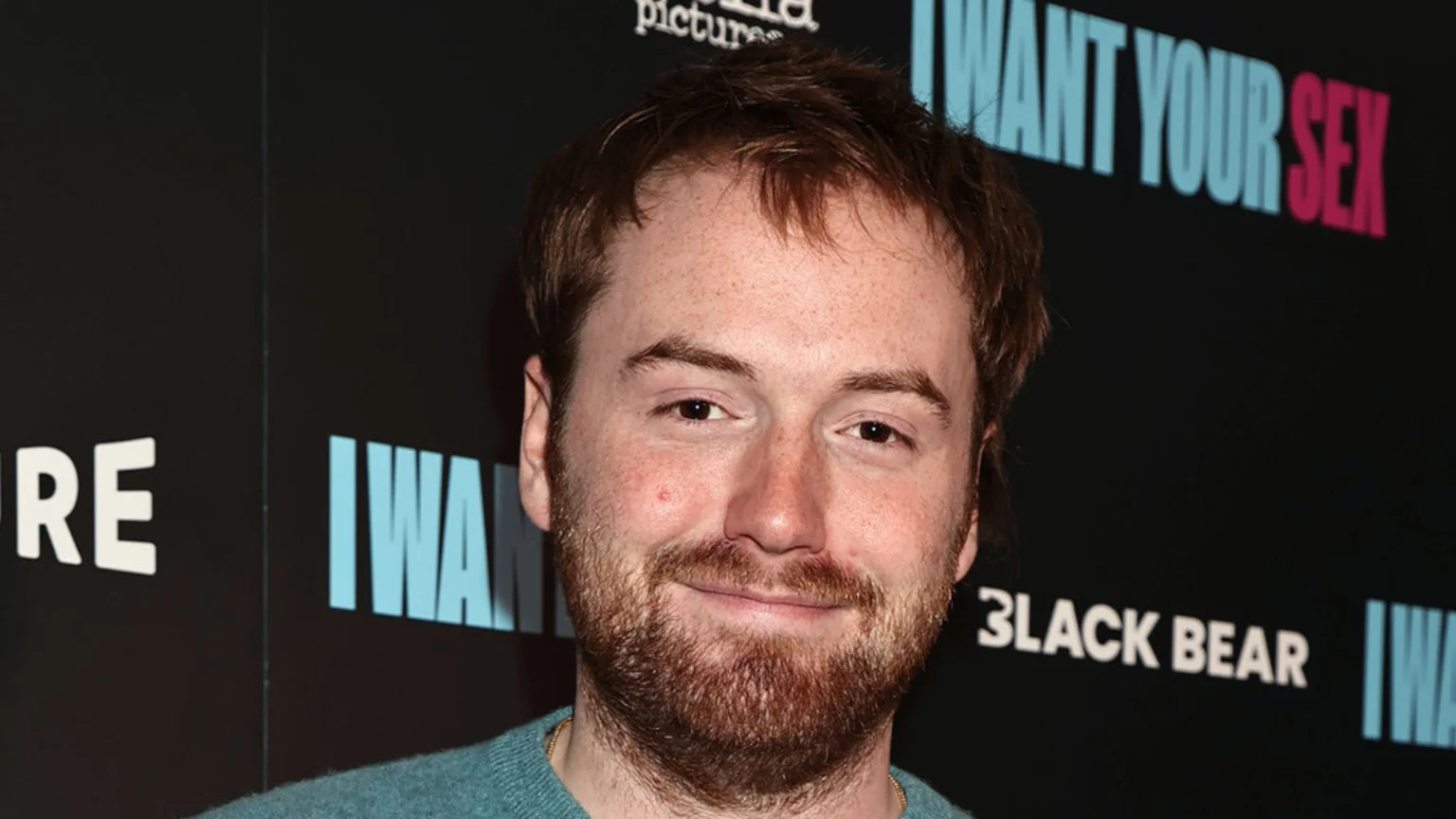 A smiling man with reddish-brown hair and a beard, wearing a teal sweater, stands in front of a dark backdrop with light blue and pink text, including "I WANT YOUR SEX" and "BLACK BEAR."