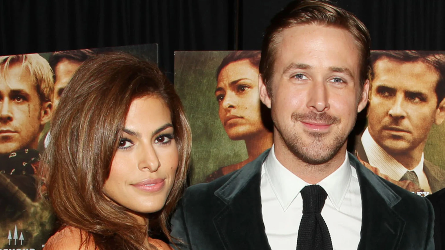 Ryan Gosling in a suit and Eva Mendes in an orange top pose together, with movie posters for "The Place Beyond the Pines" featuring themselves and Bradley Cooper visible in the background.