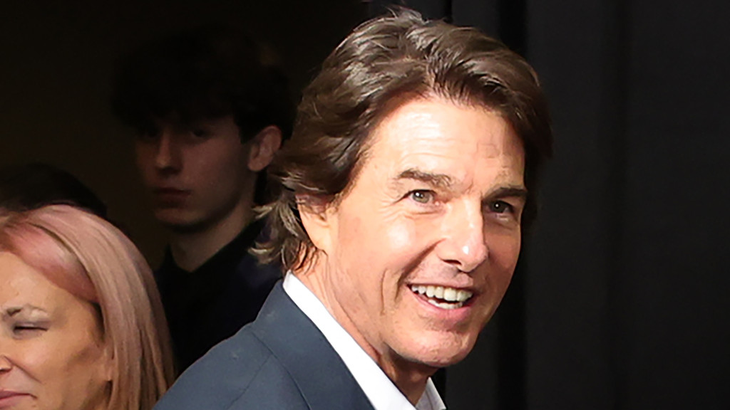 Tom Cruise smiles brightly, wearing a dark suit and white shirt, with people partially visible behind him in a dark setting.
