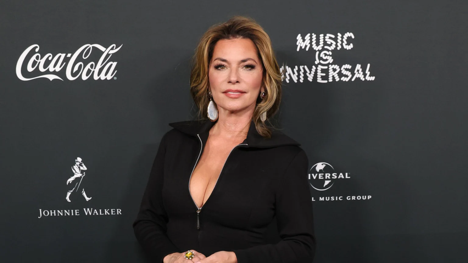 A woman with blonde hair, wearing a black zippered top and large dangle earrings, stands against a dark backdrop with logos for Coca-Cola, Johnnie Walker, Music is Universal, and Universal Music Group.