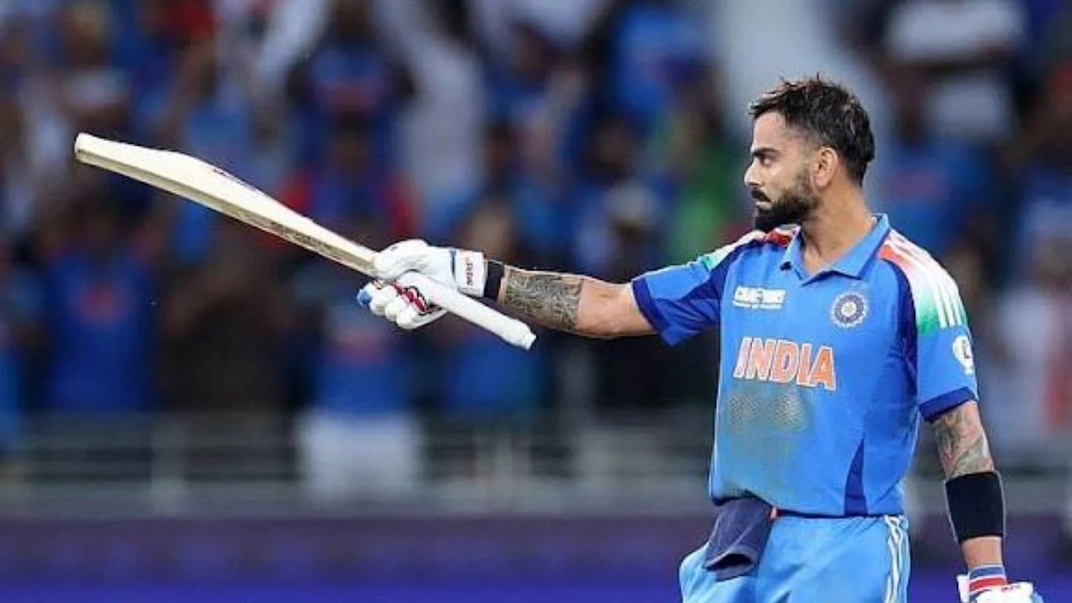 Virat Kohli in a blue Indian cricket jersey and batting gloves holds a bat, looking to his left with a blurred stadium background.