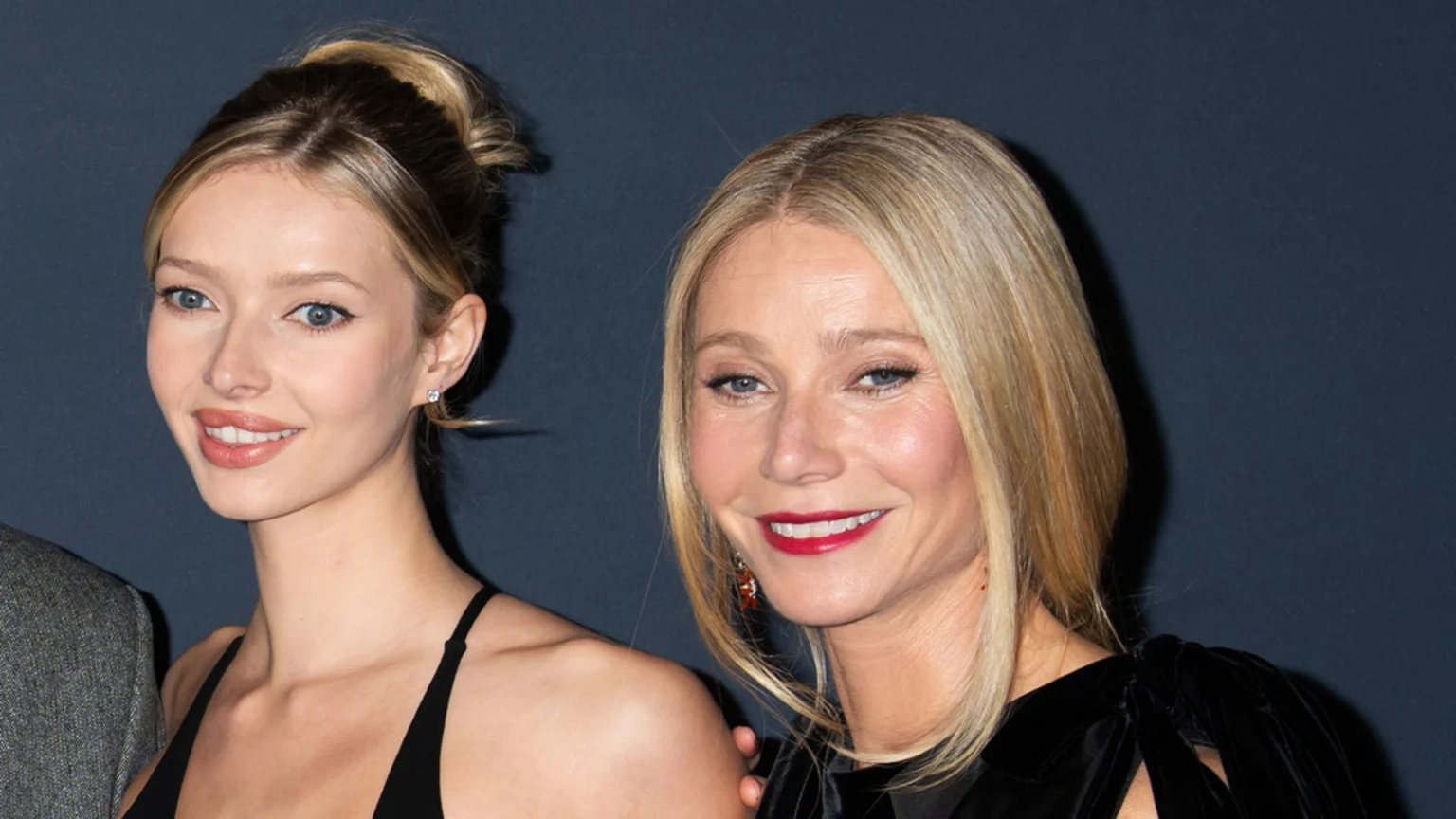 An older blonde woman in a dark top with red lipstick and red earrings smiles next to a younger blonde woman in a black strap top, against a dark blue background.