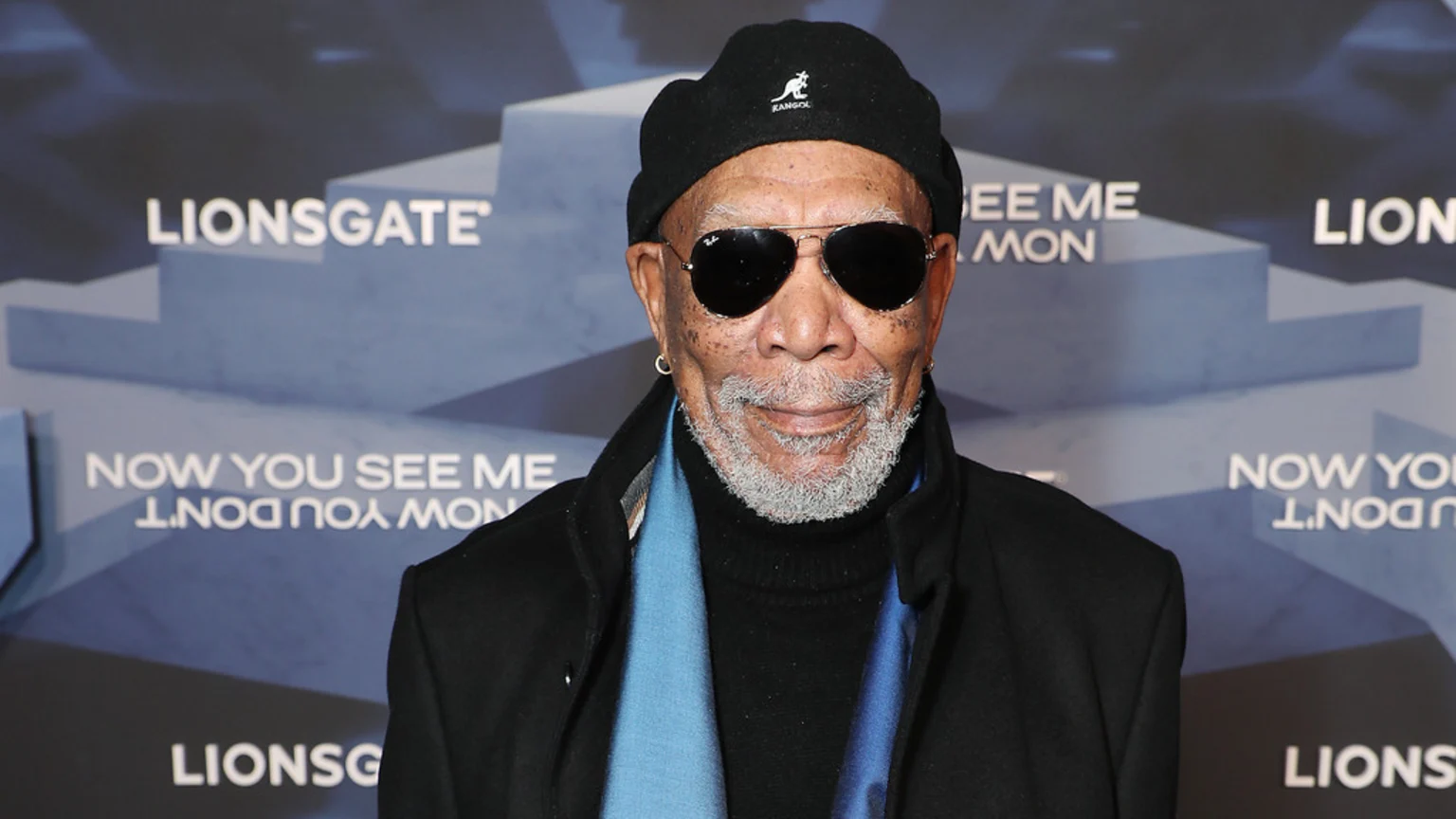 Morgan Freeman smiles, wearing a black Kangol beanie, aviator sunglasses, a black coat, and a blue scarf, against a promotional background for Lionsgate and "Now You See Me".