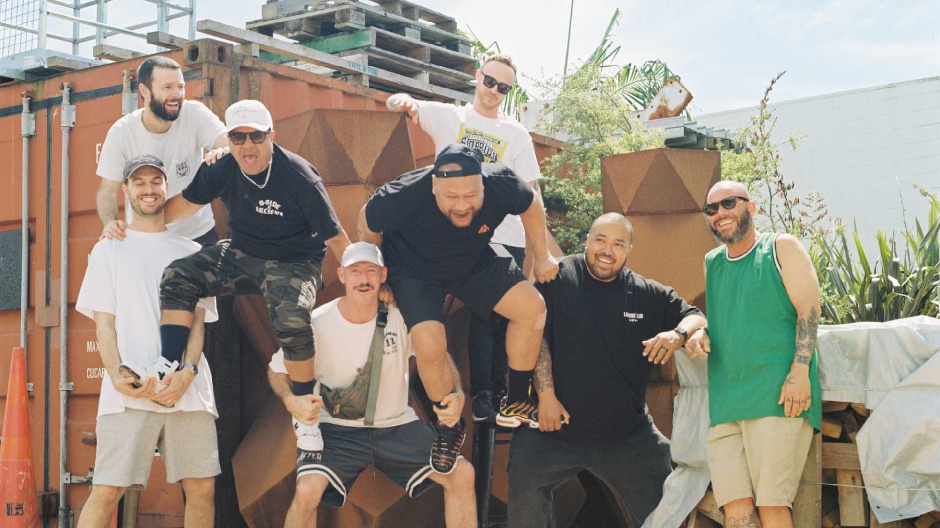 Eight men playfully stacked on each other's shoulders and backs, smiling outdoors in front of shipping containers and greenery.