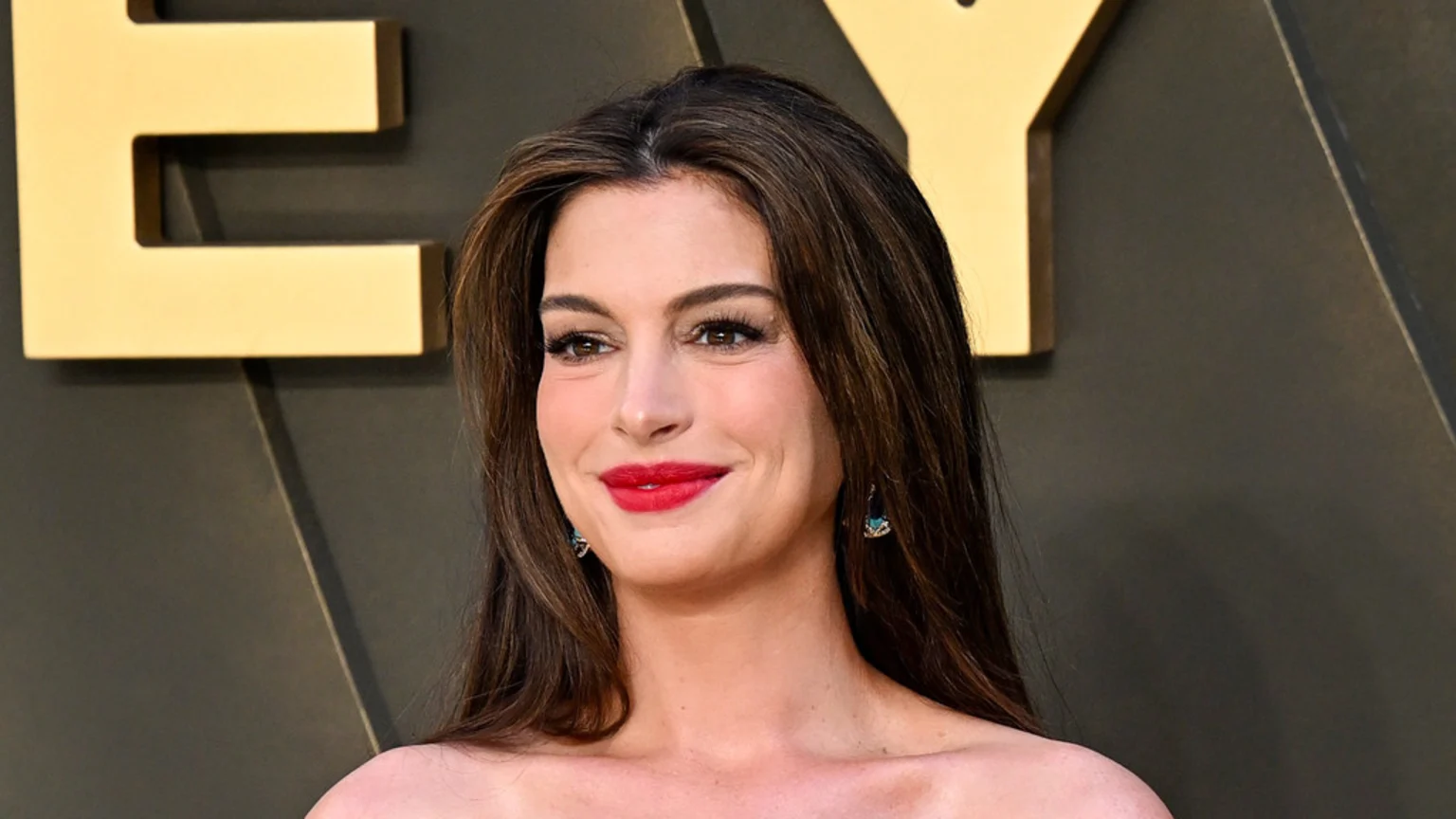 Anne Hathaway with long brown hair and red lipstick, smiling slightly and wearing turquoise earrings, against a blurred dark background with golden letters.