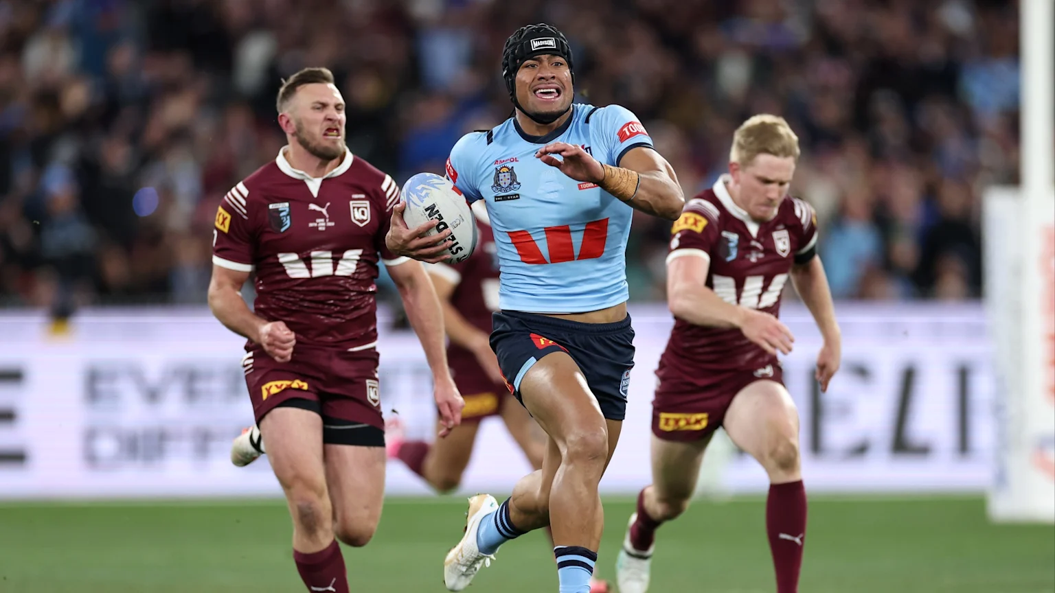 A light blue NSW rugby league player with a headguard runs with the ball on a green field, pursued by two maroon Queensland players in a brightly lit stadium.