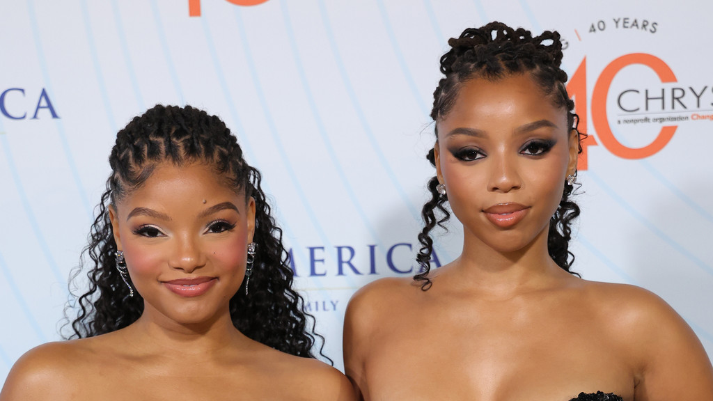 Sisters Halle and Chloe Bailey pose for a photo; Halle smiles broadly with light makeup, while Chloe has dark eye makeup and a subtle smile.