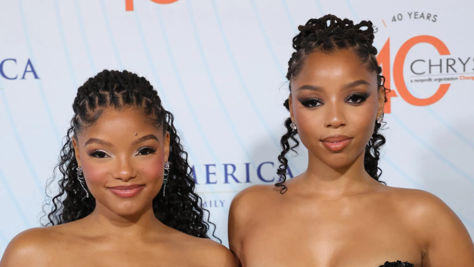 Sisters Halle and Chloe Bailey pose for a photo; Halle smiles broadly with light makeup, while Chloe has dark eye makeup and a subtle smile.