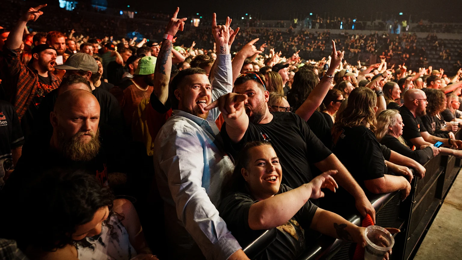 A large crowd of enthusiastic fans at a concert, many raising their hands and making rock-on gestures, with several people in the foreground smiling and pointing at the camera.