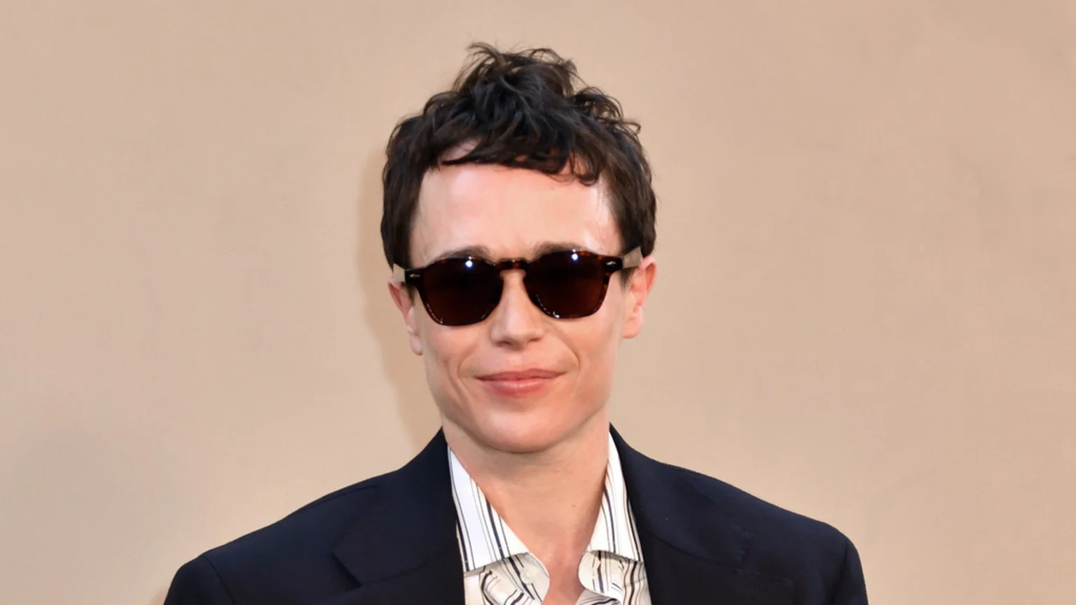 A man with short dark hair and dark sunglasses smiles faintly, wearing a dark blazer over a striped shirt, against a light tan background.