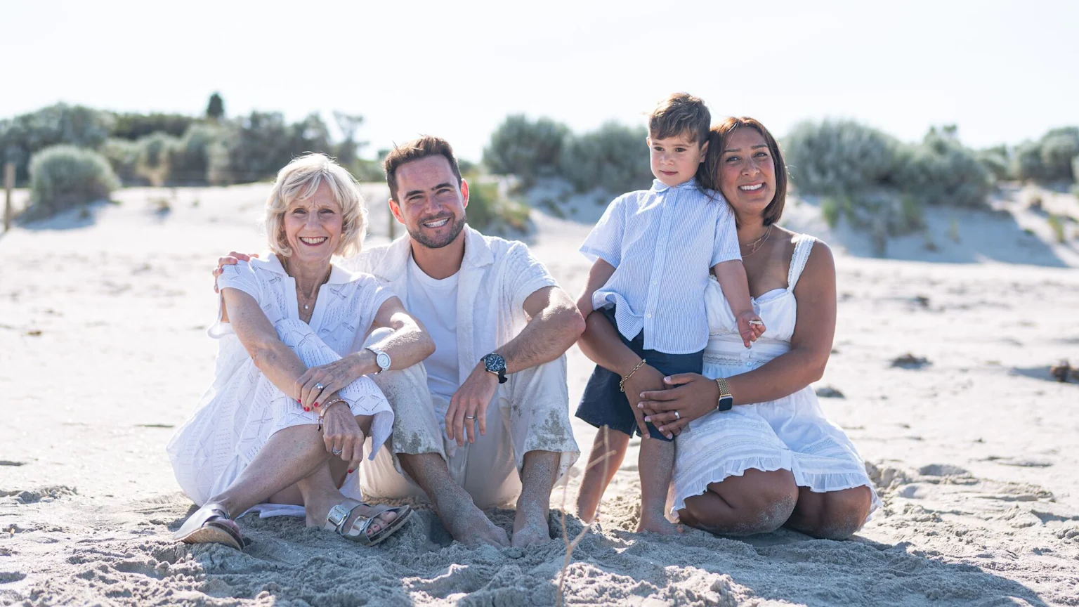 A happy family of four, including an older woman, a man, a woman, and a young boy, smiles while sitting on a sandy beach with dunes in the background.
