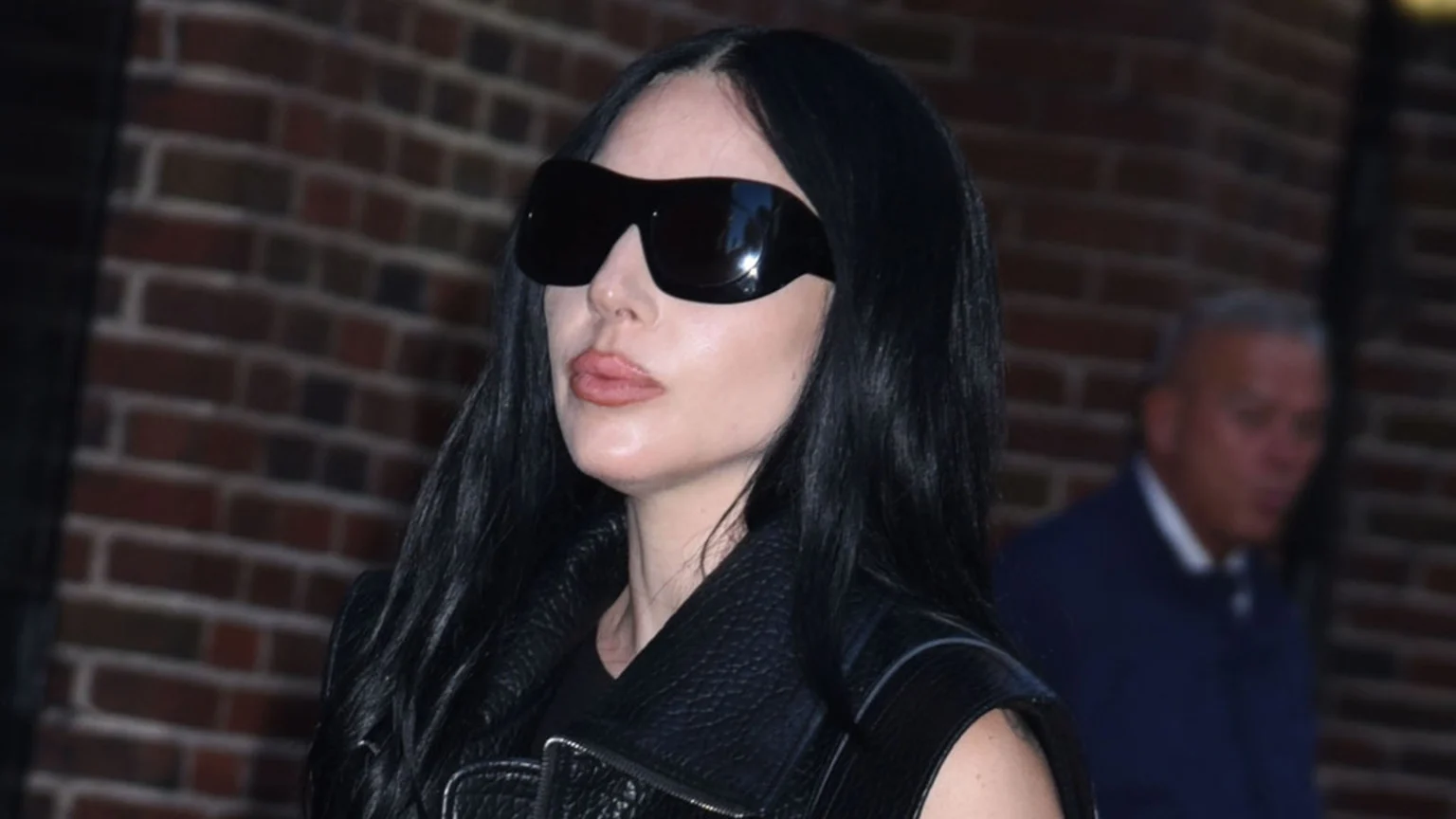 A person with long black hair, large sunglasses, and nude lipstick wears a black leather vest against a brick wall, with a blurry figure behind them.