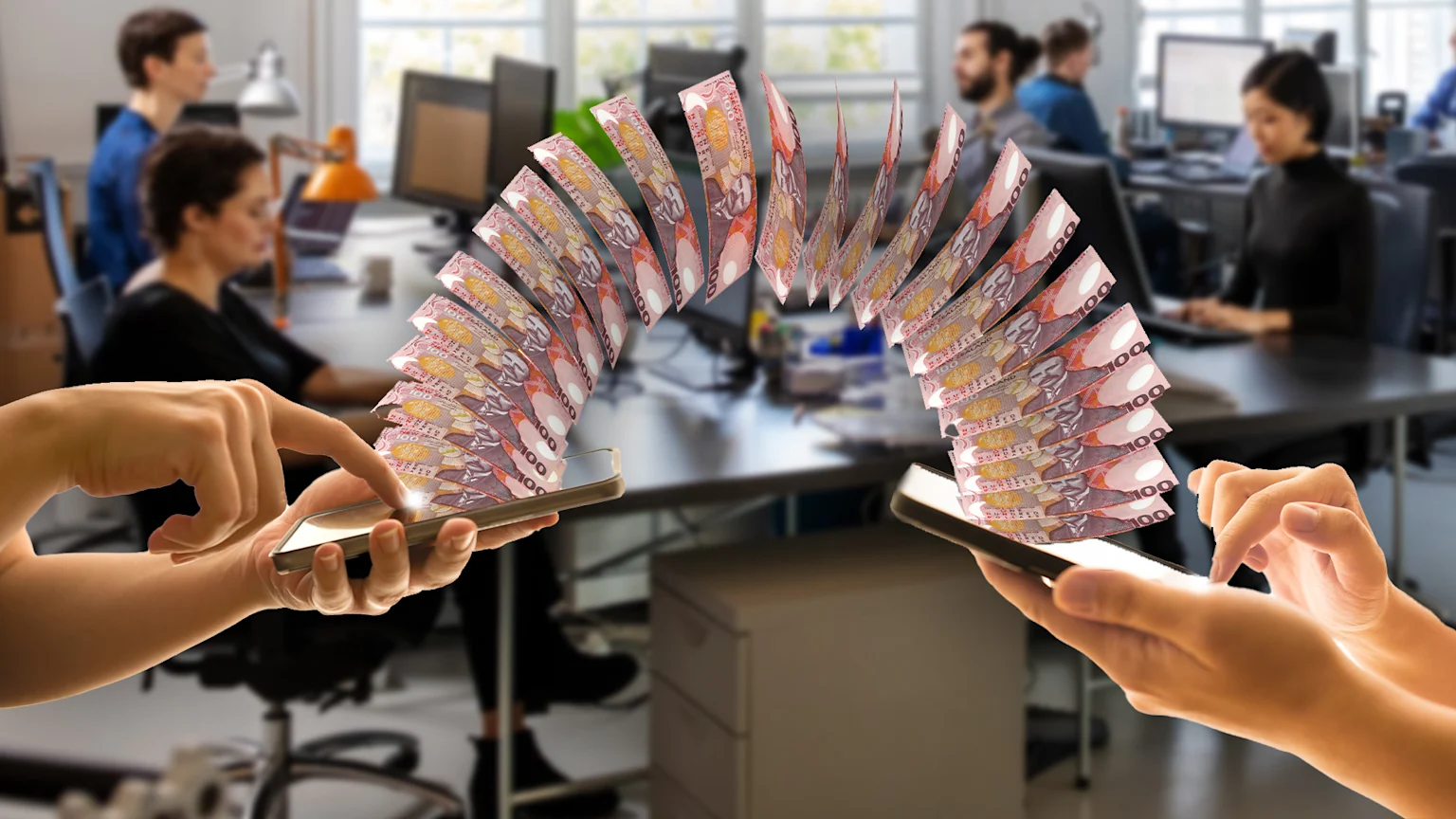 New Zealand $100 banknotes digitally arc between two smartphones held by hands, with a blurred office of workers in the background.