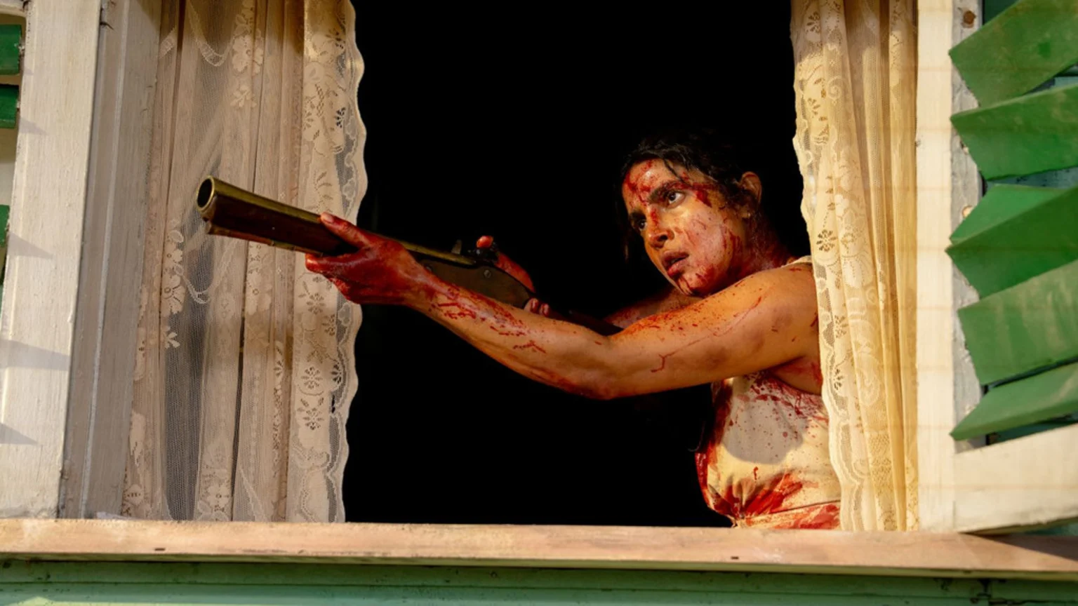 A woman, covered in blood, aims a rifle out of a window with intense focus.