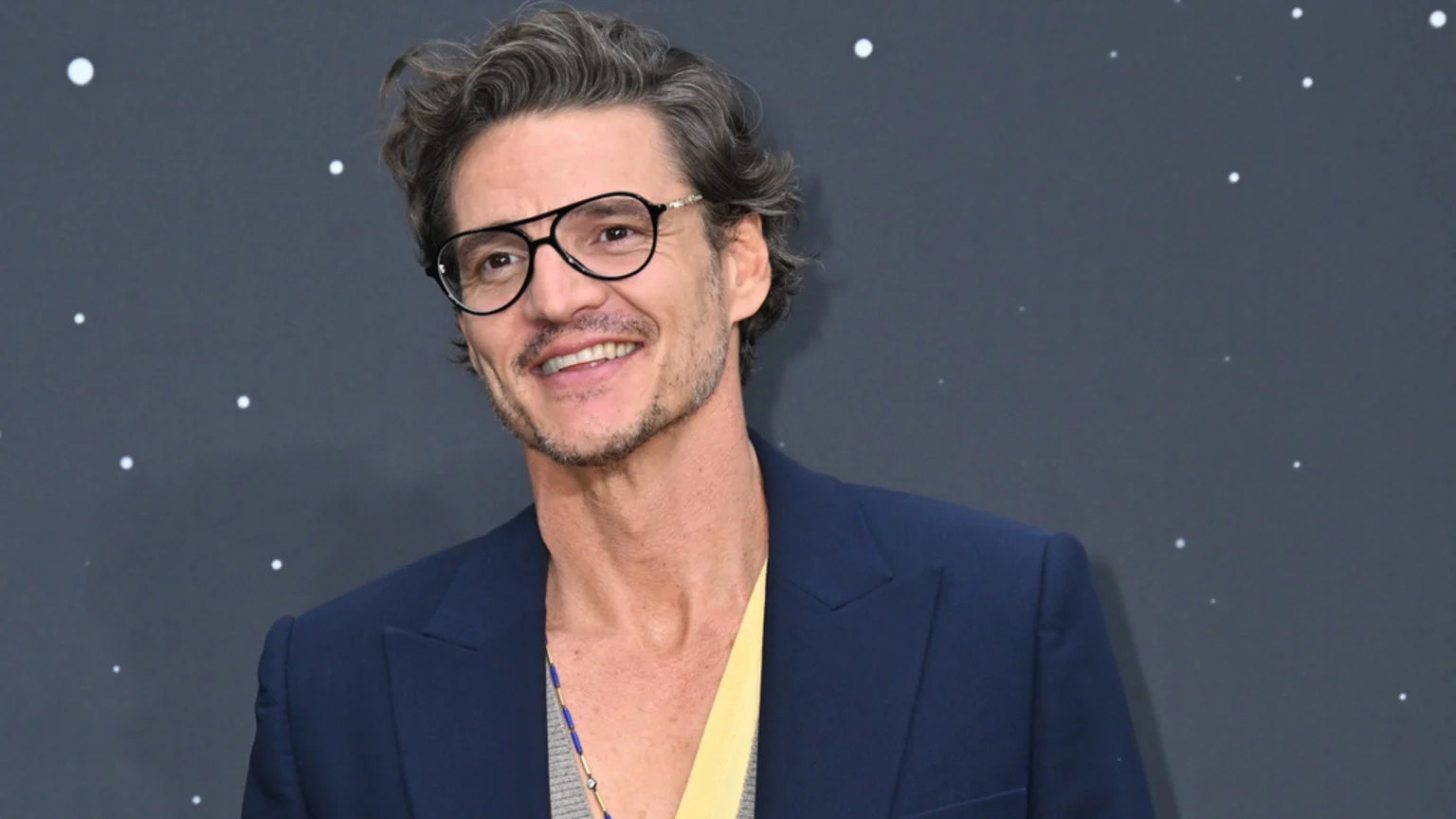 Pedro Pascal smiles, wearing glasses, a dark blue jacket, and a yellow shirt, against a dark background dotted with white specks resembling stars.