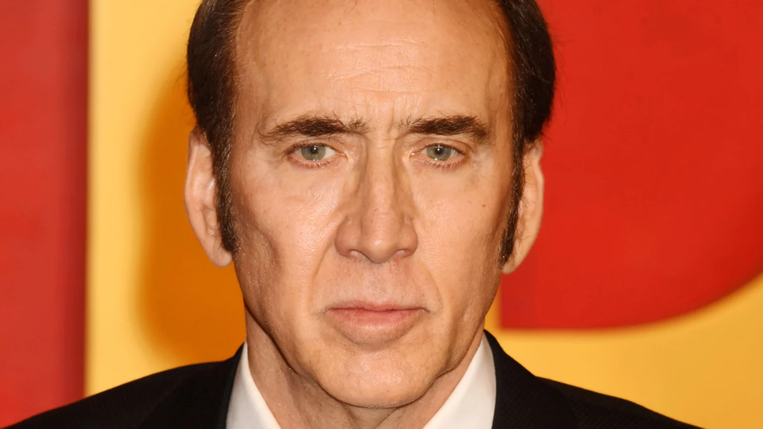 A close-up headshot of actor Nicolas Cage, looking seriously at the camera, against a blurred red and yellow background.