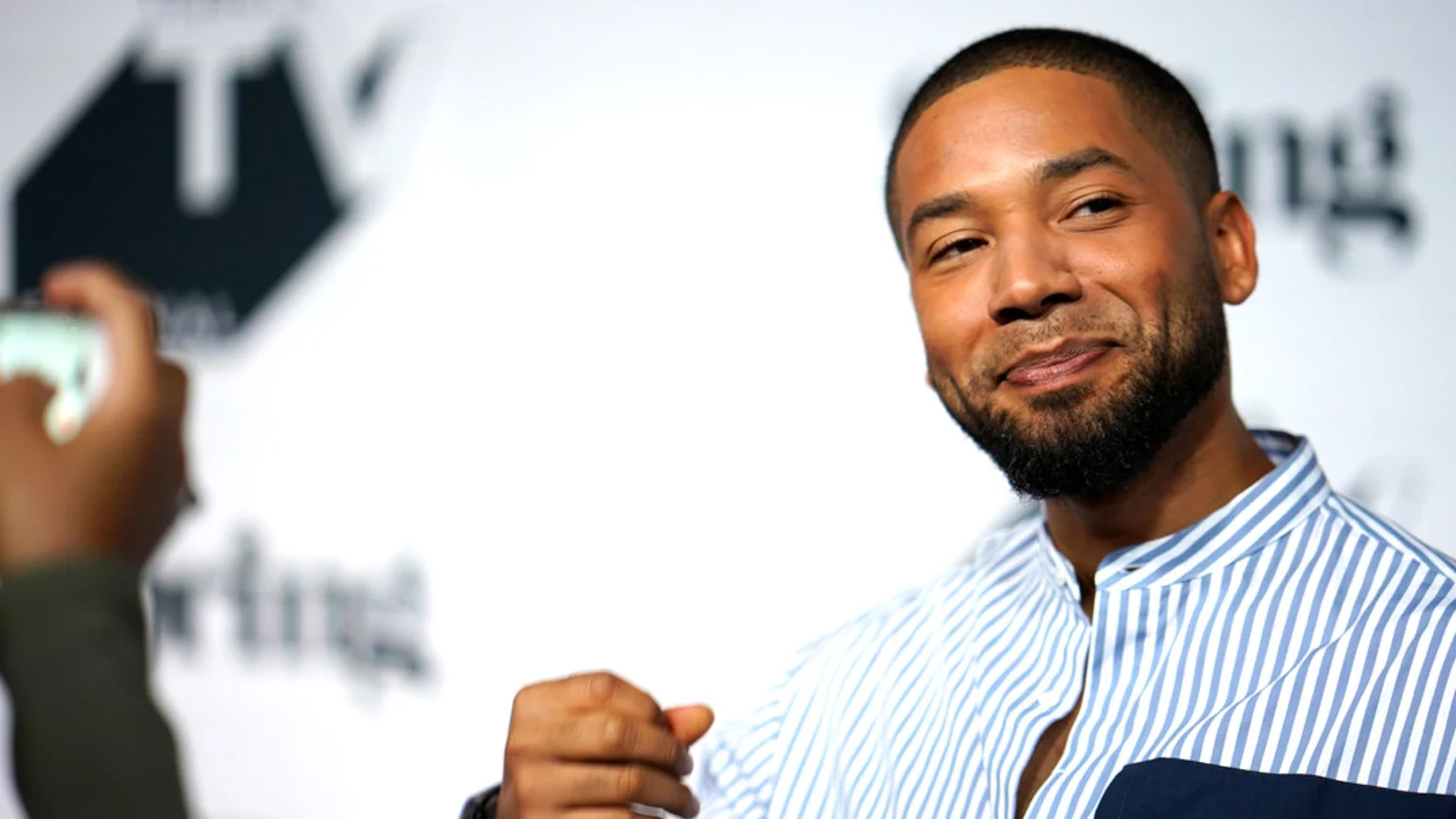 Jussie Smollett, a man with a beard and short hair, smiles slightly while wearing a blue and white striped shirt, with a blurry background and a hand holding a phone on the left.
