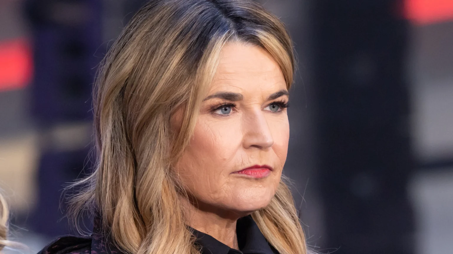 A close-up of a woman with blonde hair and dark roots, blue eyes, and visible wrinkles, wearing red lipstick and a dark shirt, looking seriously to her right.