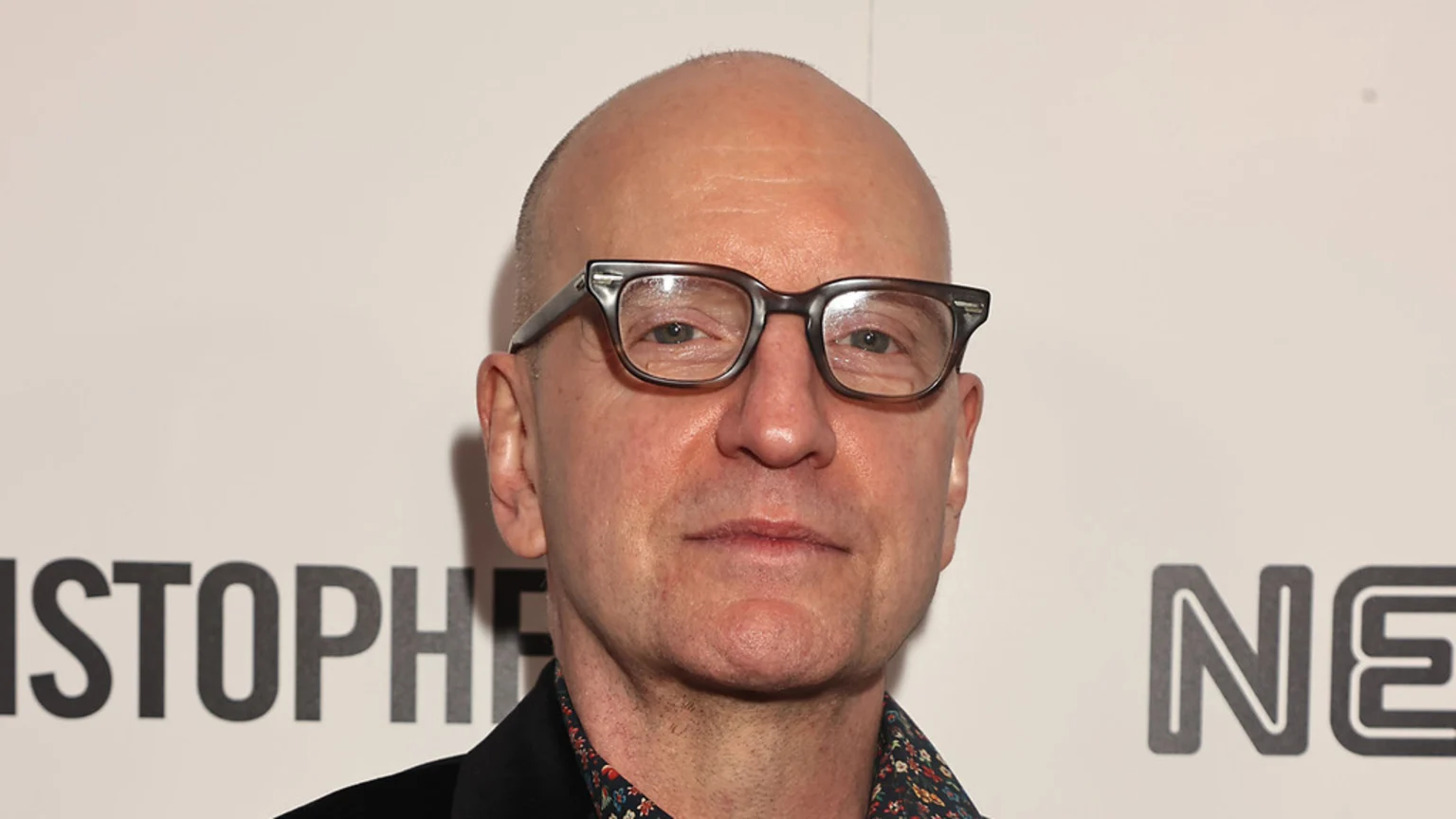 A bald man with rectangular glasses and a patterned shirt under a dark jacket looks directly at the camera against a white background with partial dark grey text visible.