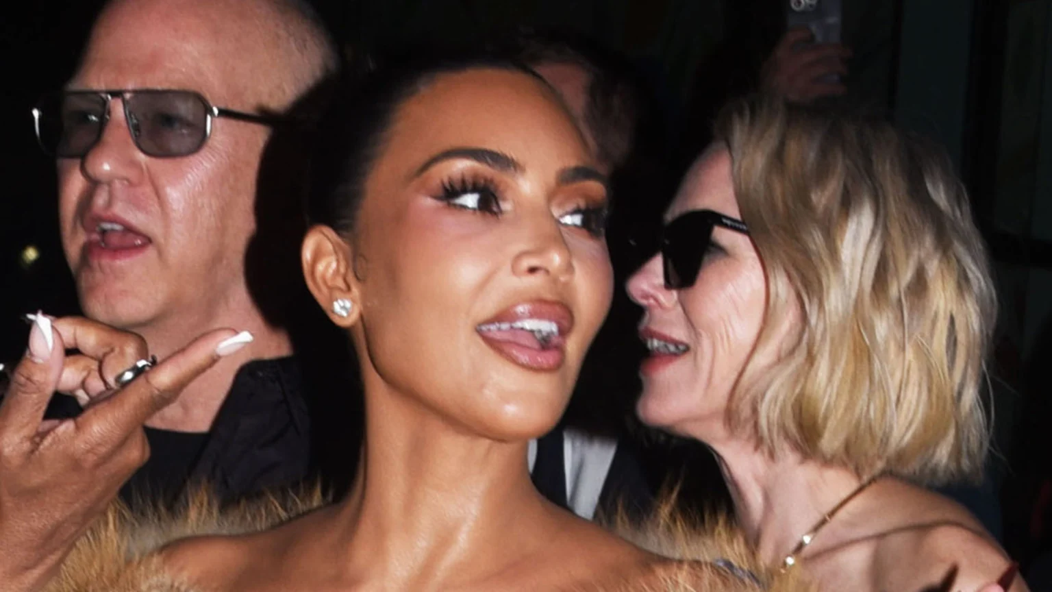 A dimly lit indoor candid photo shows Kim Kardashian with her mouth open and tongue visible, wearing a diamond stud earring and off-shoulder top with fur trim. Naomi Watts smiles beside her, wearing dark sunglasses and a gold necklace. An unknown bald man with dark sunglasses is partially visible, speaking.