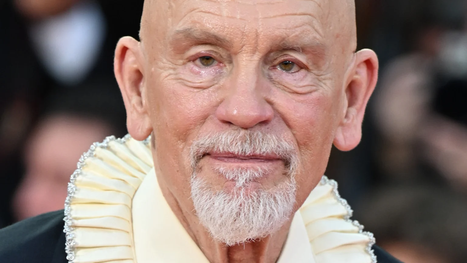 Close-up of bald John Malkovich with a white goatee, his brown eyes appearing slightly watery and reddish, wearing a cream ruffled collar with sparkling trim.