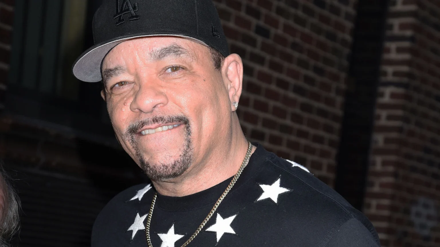 Close-up of Ice-T smiling, wearing a black LA baseball cap, a diamond earring, a gold chain, and a black shirt with white stars, against a brick wall.