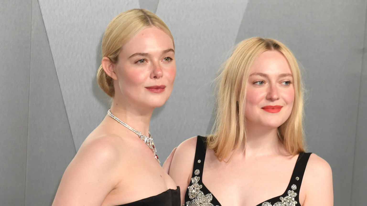 Elle Fanning, left, with hair up and a diamond necklace, stands beside Dakota Fanning, right, with loose hair and an embellished black dress, against a grey background.