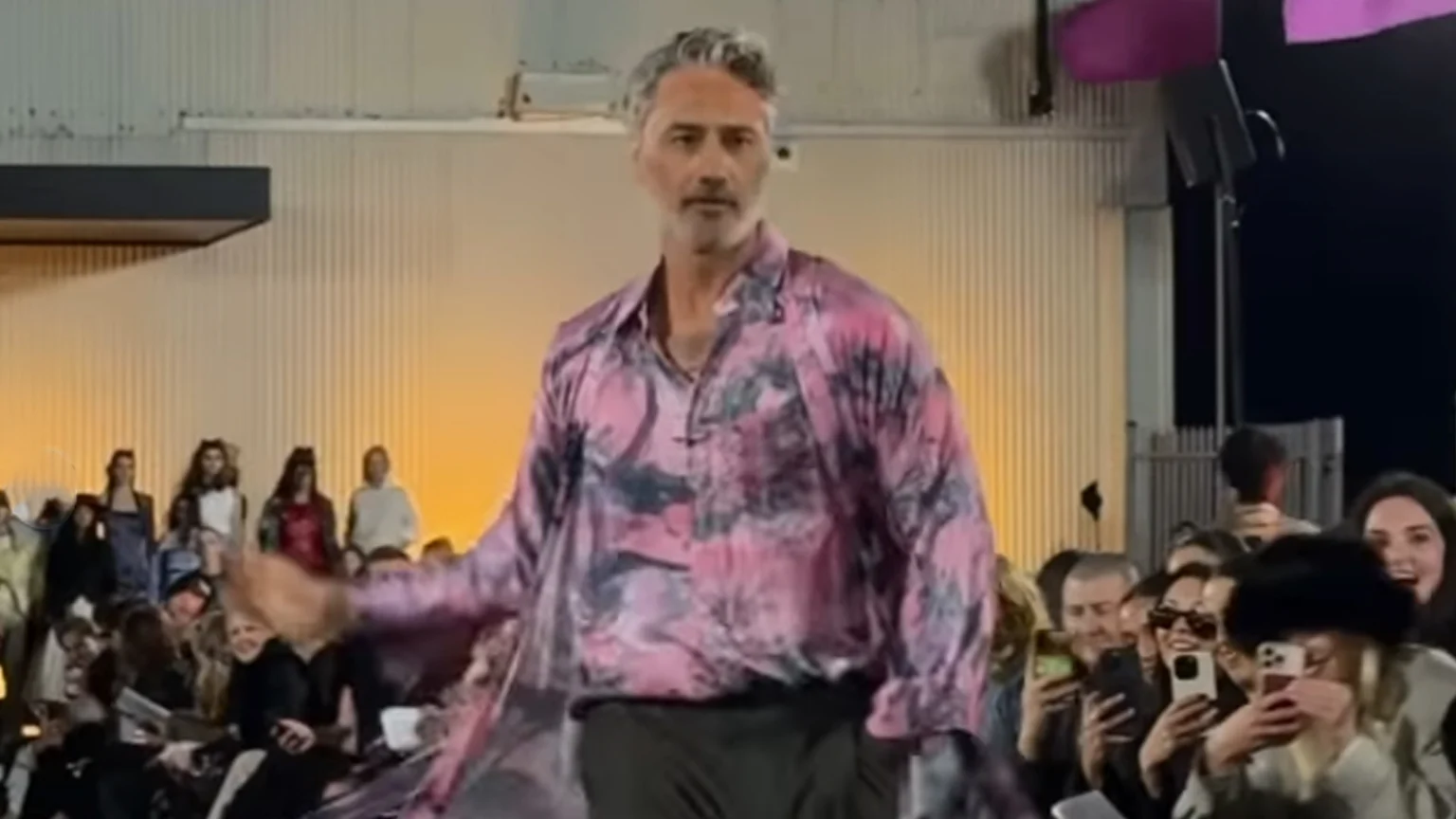 Taika Waititi walking the New Zealand Fashion Week runway outside Auckland's Shed 10 at night. He's wearing a Zambesi outfit with a silk pink and black tie dye shirt and charcoal cropped pants.