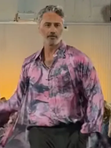 Taika Waititi walking the New Zealand Fashion Week runway outside Auckland's Shed 10 at night. He's wearing a Zambesi outfit with a silk pink and black tie dye shirt and charcoal cropped pants.